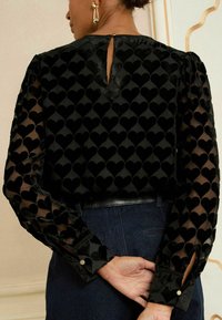 Melnā blūze ar caurspīdīgām piedurknēm, kas rotātas ar samta sirds rakstiem. Arī ar atslēgas cauruma aizmugurē un no pogas slēgšanu, un gludu auduma tekstūru.