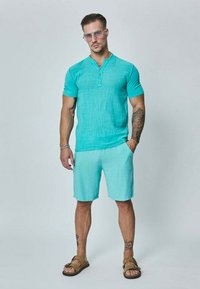 Chemise turquoise à manches courtes avec boutons et tissu texturé, associée à un short assorti. Portée avec des sandales marron et des lunettes de soleil oversize.