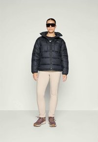 Svart vadderad jacka med hög krage, dragkedjeförslutning och stilrena sömmar. Bärs med beige åtsittande byxor och bruna vandringsskor.