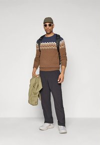 Braun- und marineblauer gemusterter Pullover, dunkle Hose, beiger Mantel, Sonnenbrille, Rucksack und Turnschuhe. Mann steht in lässiger Haltung.
