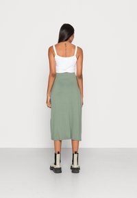 Mujer de espaldas vistiendo una camiseta sin mangas blanca, falda midi verde y botines beige con negro, con un tatuaje vertical en la espalda.