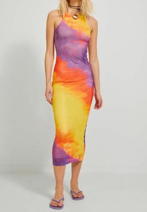 Kobieta ubrana w bezrękawową, sięgającą kostek sukienkę tie-dye w kolorach żółtym, pomarańczowym i fioletowym, z fioletowymi klapkami i naszyjnikiem z wisiorkiem w kształcie serca.