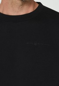 T-shirt in cotone nero con scollo rotondo e una texture morbida. Logo "SAIL RACING" stampato in una tonalità grigia sobria sul davanti.