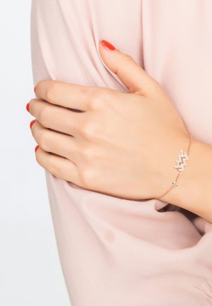 Gold-tone bracelet with a zigzag design, featuring clear stones and a small round accent. Worn on a hand against a soft pink fabric.