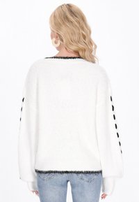 Weißer Pullover mit lockerer Passform, strukturiertem Strick, schwarzen Akzenten an Kragen und Ärmeln sowie dekorativen schwarzen Knöpfen an den Ärmeln.