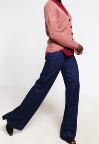 Lyserød oversized cardigan med blød tekstur, lukket med knapper, båret over en mørkerød skjorte, kombineret med mørkeblå vidde-bukser.