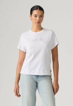Mujer con cabello oscuro recogido lleva una camiseta blanca de Levi's y vaqueros azul claro, de pie frente a un fondo liso de color gris claro.