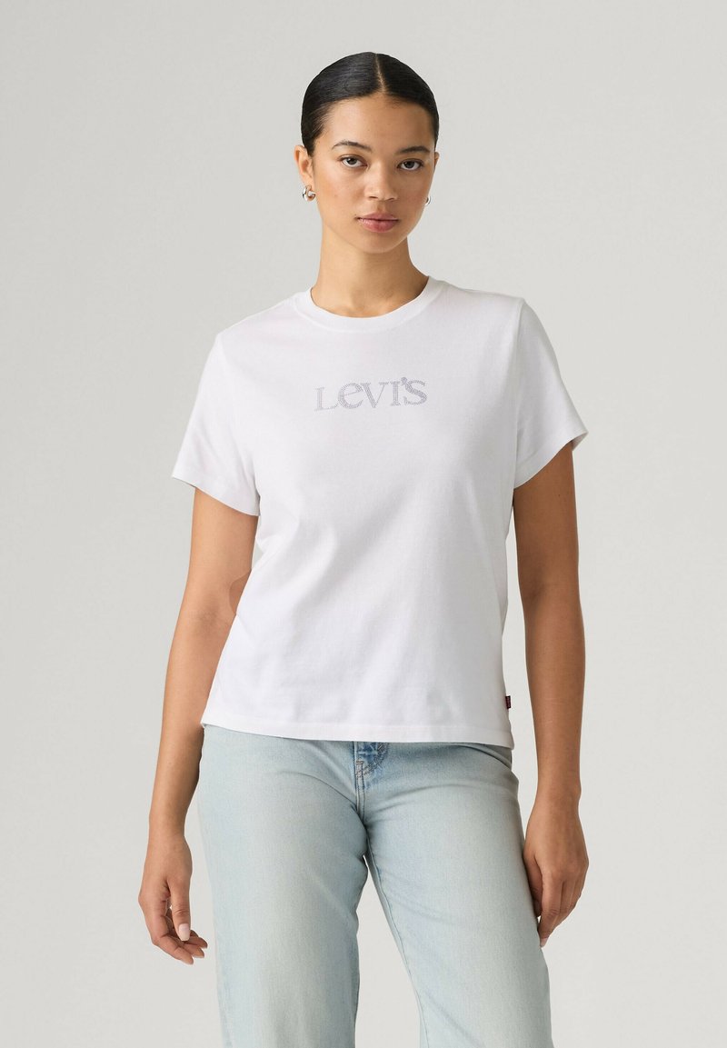 Femme aux cheveux foncés attachés en arrière porte un t-shirt blanc Levi's et un jean bleu clair, debout devant un fond gris clair uni.
