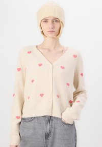 Beež kardigan roosa südame mustriga, v-kaelusega, nööbitava kinnitusega ja soonikvarrukkidega. Kombineeritud beeži kudumisega beanie'ga.