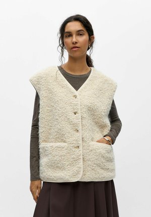 Femme portant un gilet sans manches texturé couleur crème sur une chemise à manches longues foncée et une jupe sombre, une main dans la poche et une expression neutre.