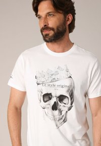 T-shirt en coton blanc avec un graphisme de skull noir surmonté d'une couronne et le texte "Le style original 1974" en dessous. Manches courtes.