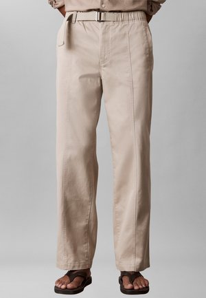 Beżowe spodnie o prostych nogawkach z szlufkami na pasek i bocznymi kieszeniami, noszone z brązowymi sandałami na szarym tle.