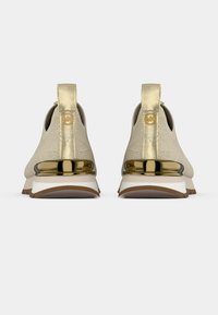 Beiget urheilukengät, joissa on teksturoitu päällinen, kullanvärisiä yksityiskohtia, pehmustettu kanta ja valkoinen välipohja. Kantapäässä on logo.