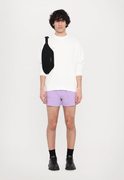 Sweatshirt blanc, shorts violets, sac bandoulière noir. Le modèle porte des chaussettes noires et des baskets. Design simple, tissu décontracté, style minimaliste.