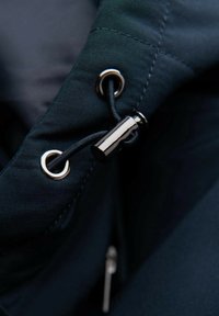 Närbild av ett mörkt tyg med en silverfärgad ögla och svart snöre. Innehåller en blank svart justeringsknapp och synliga sömnadsdetaljer.