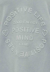 Tessuto azzurro chiaro con testo in rilievo che recita "POSITIVE LIFE GOOD ENERGY POSITIVE MIND CLUB" con cuori accentuati, la texture è morbida.