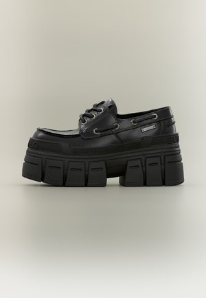 Chaussure à plateforme noire épaisse avec laçage sur le devant, semelle épaisse et robuste, et détails de marque le long de la semelle et sur le côté.