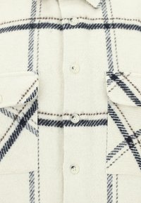 Biała flanelowa koszula w czarno-brązowy wzór w kratę, z podwójnymi kieszeniami na piersiach i sześcioma guzikami z przodu. Teksturowana tkanina.