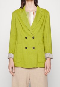 Donna che indossa un blazer doppiopetto verde lime con bottoni neri, polsini di camicia stampati, pantaloni beige, in piedi contro uno sfondo bianco.