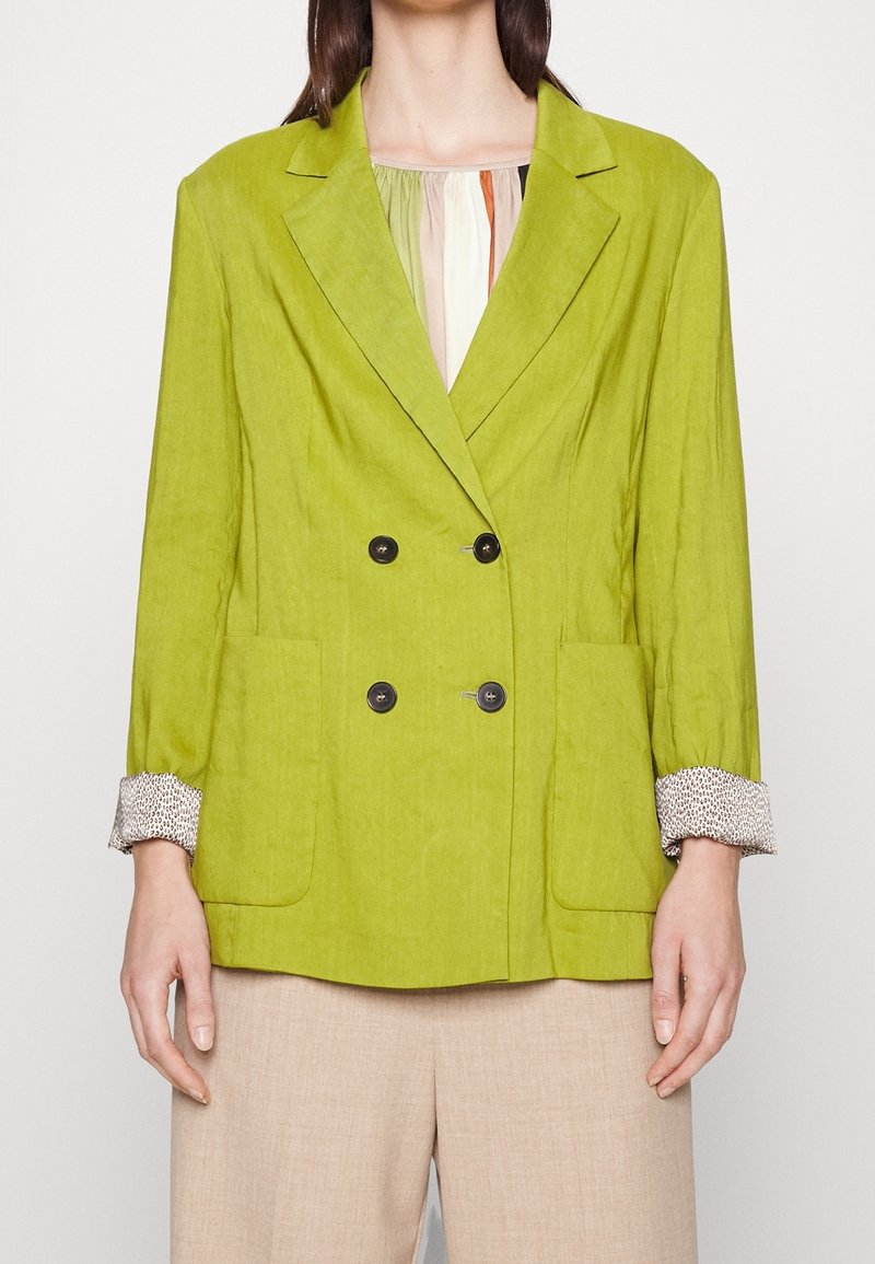 Donna che indossa un blazer doppiopetto verde lime con bottoni neri, polsini di camicia stampati, pantaloni beige, in piedi contro uno sfondo bianco.