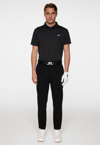 Svart kortärmad pikétröja med en liten vit logotyp, kombinerad med svarta byxor, ett svart bälte och en vit golfhandske, bärs tillsammans med vita sneakers.