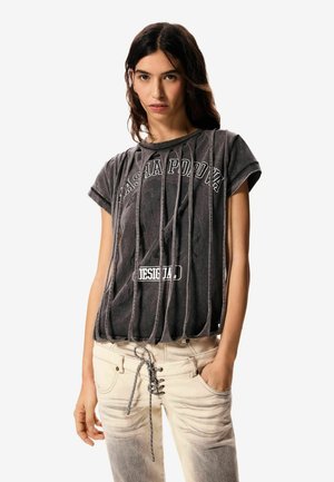 Kvinde med langt mørkt hår iført sort, slidt t-shirt med grafik og lyse, falmede jeans med snørelukning foran, hænderne i lommerne.