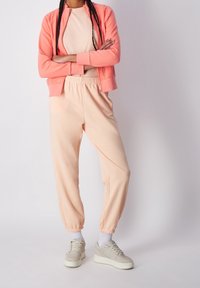 Ensemble de survêtement rose clair comprenant un hoodie zippé avec des manches côtelées et un pantalon de jogging fuselé assorti avec une taille élastique, porté avec des baskets.