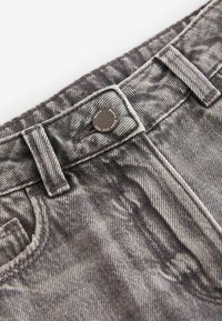 Jeans in denim grigio con una finitura sbiadita e testurizzata, dotati di chiusura a bottoni e passanti per cintura. La cucitura è visibile lungo i bordi.