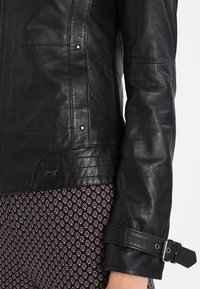 Gros plan d'une manche de veste en cuir noire avec une bride à boucle sur un pantalon à motifs sur un fond clair.