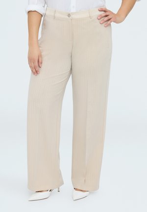 Kvinde iført beige bukser med vide ben og hvide sko med høje hæle, stående med den ene hånd på hoften og den anden afslappet ned langs siden.