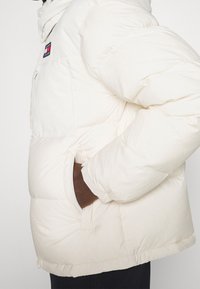 Personne portant une doudoune blanche avec une main dans la poche latérale et un écusson logo Tommy Jeans sur la poitrine.