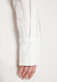 Chemise blanche à boutonnière avec un poignet ajusté comportant trois boutons blancs, un tissu lisse et un design de manche structuré. Style minimaliste.