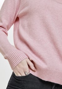 Jasnoróżowy sweter dziergany z prążkowanymi mankietami i dołem, o miękkiej fakturze, z ręką swobodnie włożoną w kieszeń.