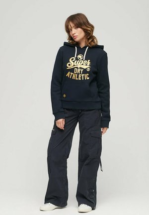 Dunkelblauer Baumwollhoodie mit goldfarbener Grafikaschrift, Fronttasche und Kordelzugkapuze, kombiniert mit locker sitzenden dunklen Cargo-Hosen und weißen Turnschuhen.