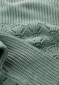 Tissu en maille texturée vert pâle ; présente un bord côtelé et des motifs ajourés complexes avec un tissage doux et dense.