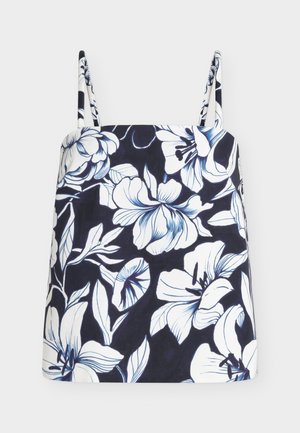 Femme aux cheveux blonds courts portant un haut floral noir et blanc, un pantalon blanc, des sandales blanches à talons, des bijoux en or et tenant un petit sac à main bleu.