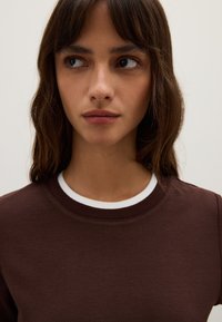 Felpa marrone con collo a girocollo bianco, maniche corte e una texture liscia, senza motivi o grafiche visibili.