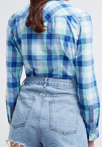 Camicia a quadri in tonalità di blu e verde, con maniche lunghe, design con bottoni e un tessuto leggero. Abbinata a pantaloni corti di denim.