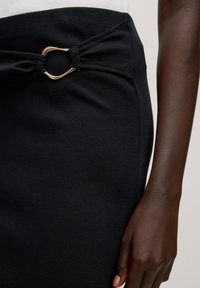 Schwarzer gerippter Rock mit einem geknoteten Design an der Taille und einem goldenen Metallakzent. Glatte Textur mit einer engen Silhouette.