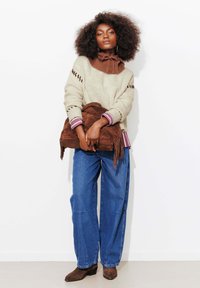 Pull en maille crème et marron avec détail nœud, assorti à un jean large bleu foncé et un sac à main en daim marron avec des accents de franges.