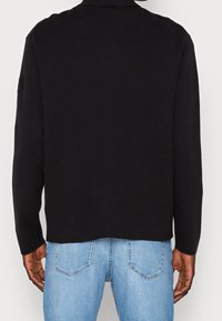 Svart långärmad topp med krage, tillverkad av mjukt tyg. Har en rak skärning och diskret sömdetalj på baksidan. Bärs över ljusblå jeans.
