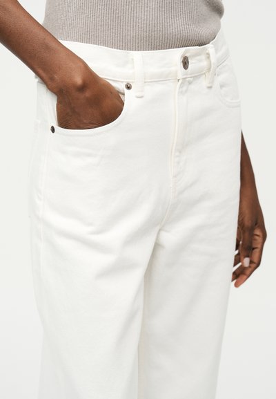 Άτομο που φοράει λευκό παντελόνι με το ένα χέρι στην μπροστινή τσέπη και μπεζ ριμπ τοπ μέσα στο παντελόνι.