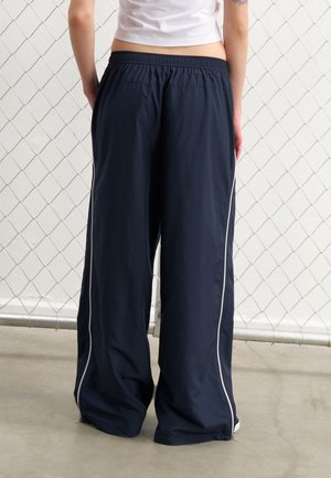 Osoba w luźnych granatowych spodniach z białymi paskami po bokach, stojąca tyłem do ogrodzenia z siatki.