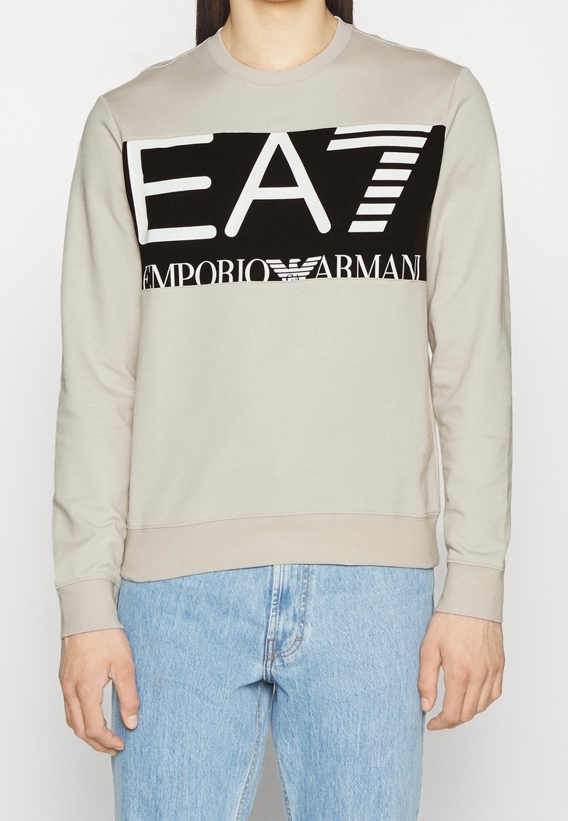 Ljusgrå sweatshirt med svart panel som har "EA7 EMPORIO ARMANI"-logotyp i vitt. Långärmad och med rund halsringning. Avslappnad passform.