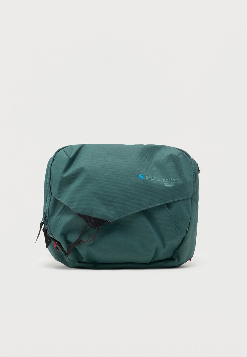 Πράσινη υφασμάτινη τσάντα μέσης με μπροστινή τσέπη με φερμουάρ στο καπάκι, μαύρα κορδόνια και μικρό μπλε λογότυπο στο μπροστινό μέρος.