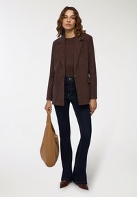 Brązowy żakiet z korduroju z pojedynczym zapięciem, noszony na brązowej bluzce i ciemnych dżinsach w stylu dzwonów. Trzyma beżową torbę o gładkiej fakturze i prostym designie.