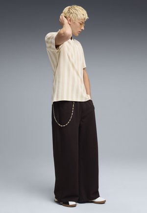 Młoda osoba z krótkimi blond włosami, ubrana w beżową koszulę w paski, szerokie czarne spodnie z łańcuchem, białe tenisówki i okulary z bursztynowymi szkłami, stojąca bokiem.