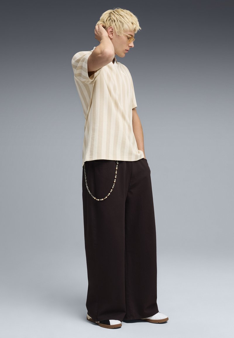 Persona joven con cabello corto y rubio, vistiendo una camisa beige a rayas, pantalones anchos negros con cadena, zapatillas blancas y gafas con cristales color ámbar, de pie de lado.