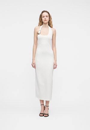 Femeie purtând o rochie lungă albă cu decolteu halter, cu o broșă circulară mare pe breteaua stângă și sandale negre cu toc înalt și barete.