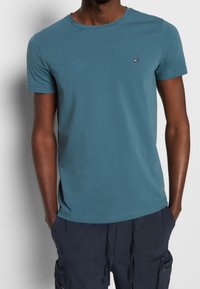 Krátký rukáv, tričko s kulatým výstřihem v modrozelené bavlně. Na hrudi se nachází malé logo a materiál je hladký s rovnoměrným povrchem.